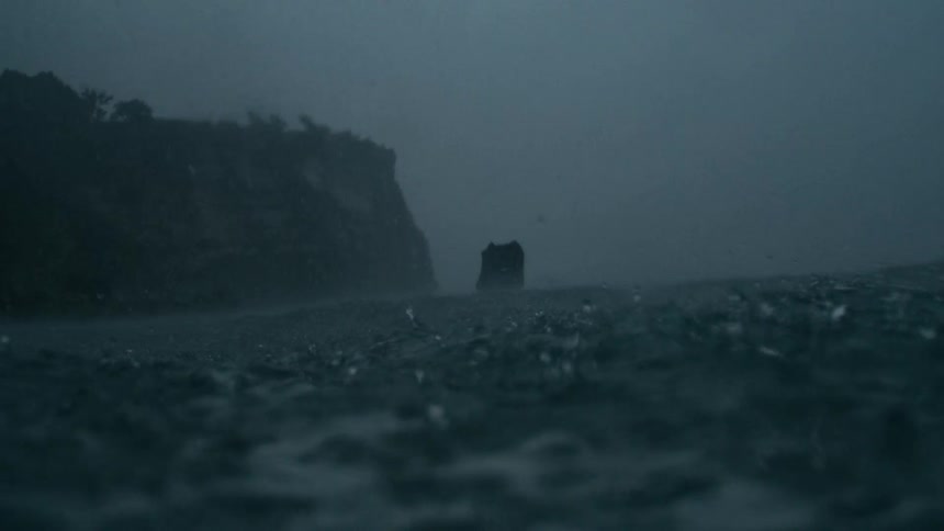 Rain falls over a fogshrouded ocean in the foreground with a distant shoreline and towering cliff partially obscured in the mist