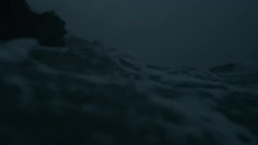 A wave crashes against jagged rocks beneath a dark stormy sky with foamy water swirling at the cliff base