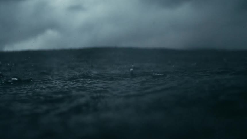 An underwater perspective with the camera partially submerged the surface of the water is choppy with ripples and splashes indicating rain falling or a strong current against a background of dark cloudy skies