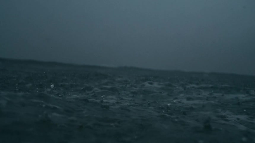 The video shows heavy rainfall on a dark body of water the water surface is agitated with numerous ripples and splashes and the background is a blurred dark horizon under a grey overcast sky