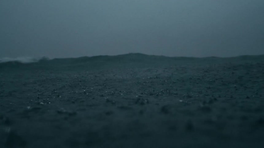 An underwater perspective of a shallow sandy seabed with numerous small dark particles or debris being stirred up the water is murky and small bubbles are visible on the surface suggesting a dynamic disturbed environment