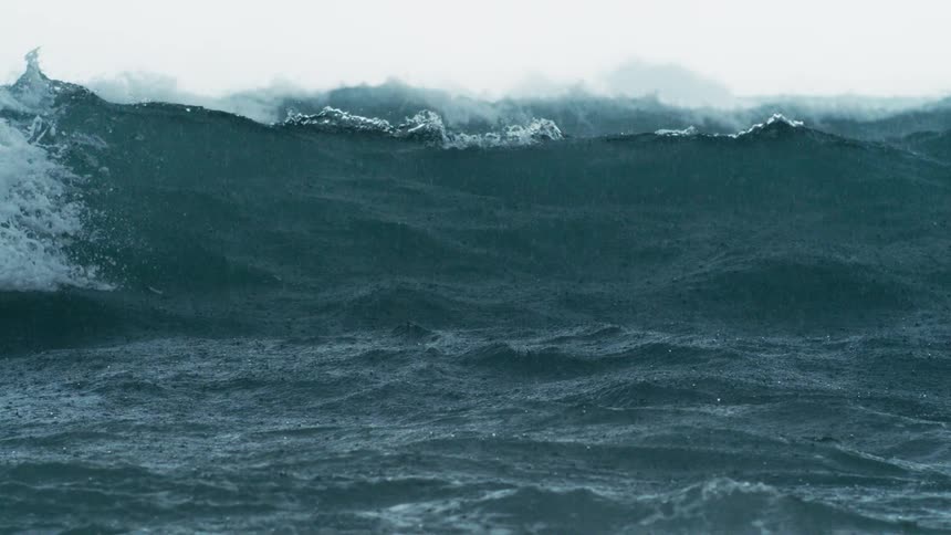 A large ocean wave surges through rainstreaked air with mist and droplets scattering across its dark choppy surface
