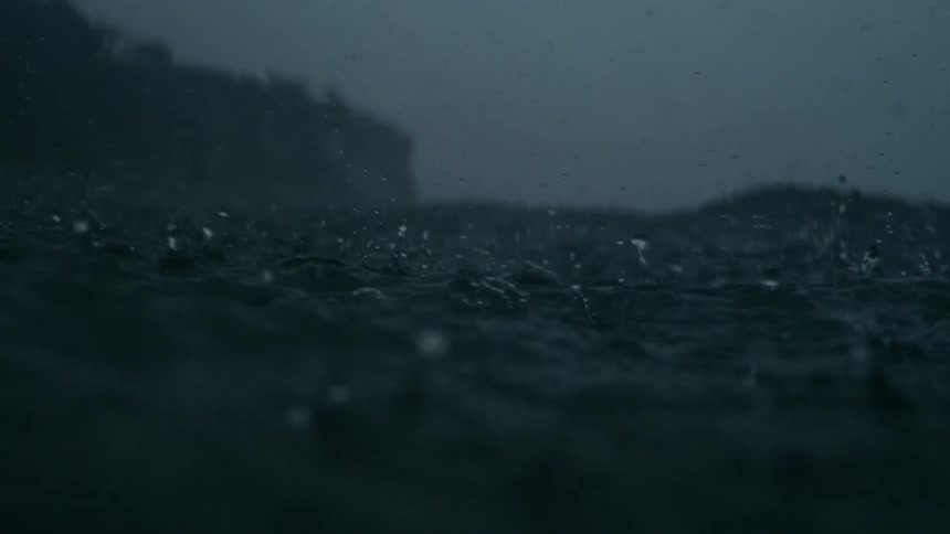 Rain falls over a fog shrouded ocean in the foreground with a distant shoreline and towering cliff partially obscured in the mist