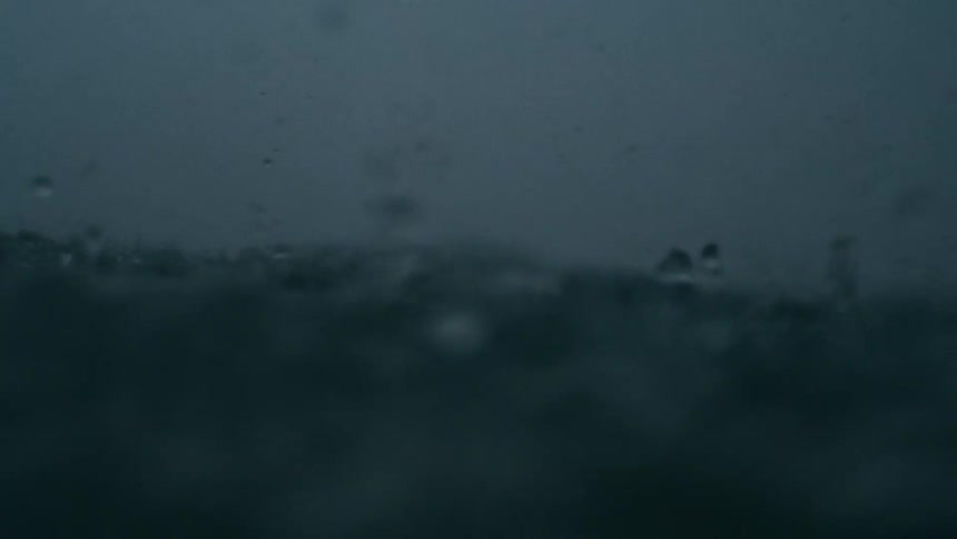 An underwater perspective where the camera is partially submerged with the surface of the water agitated by raindrops or splashing the background appears to be dark and indistinct suggesting a cloudy sky or heavy overcast conditions above the water