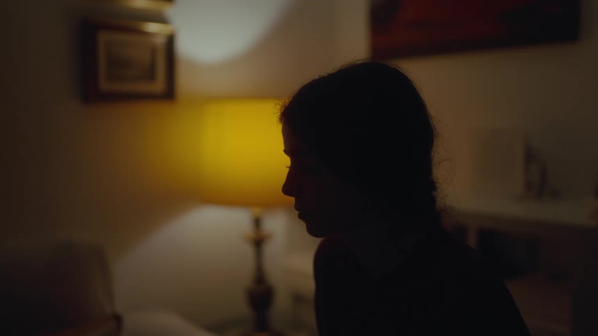 A young woman is sitting in her bedroom at night with a bedside lamp faintly illuminating the room