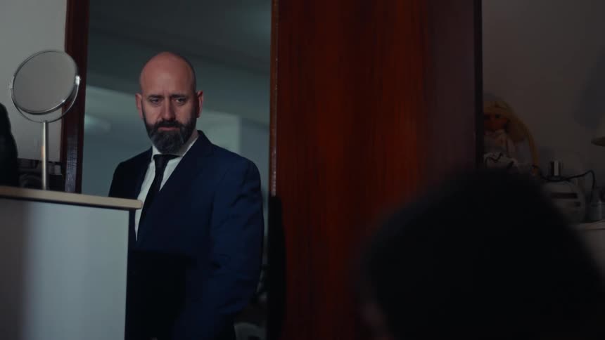 A suited person stands indoors near a mirror and wooden door casting a wistful glance toward someone in the room before quietly closing the door to leave
