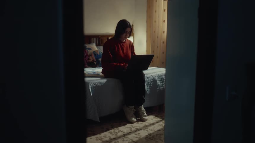 A young woman walks into a room at midday and operates her laptop while sitting on the bed