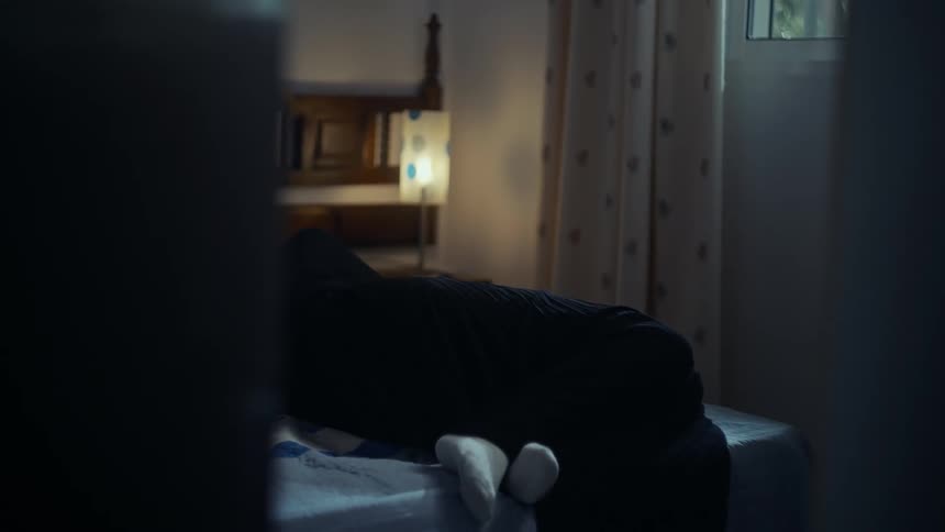 A woman sits and leans back on a bed looking sad in a dimly lit room