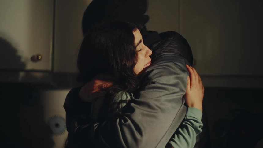 A father in a cap embraces his daughter in a dim kitchen her arms wrapped tightly around his neck in a moment of quiet closeness