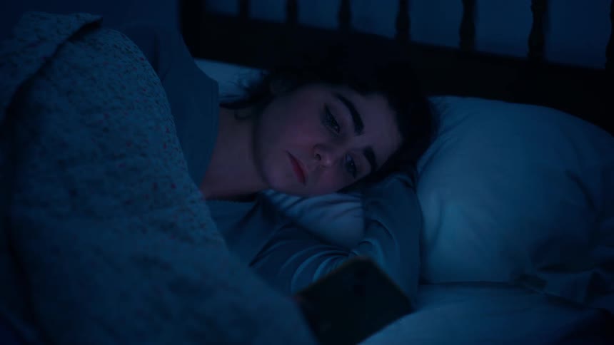 A woman lies in bed holding a smartphone partially covered by a blanket with pillows stacked behind wearing a sad expression