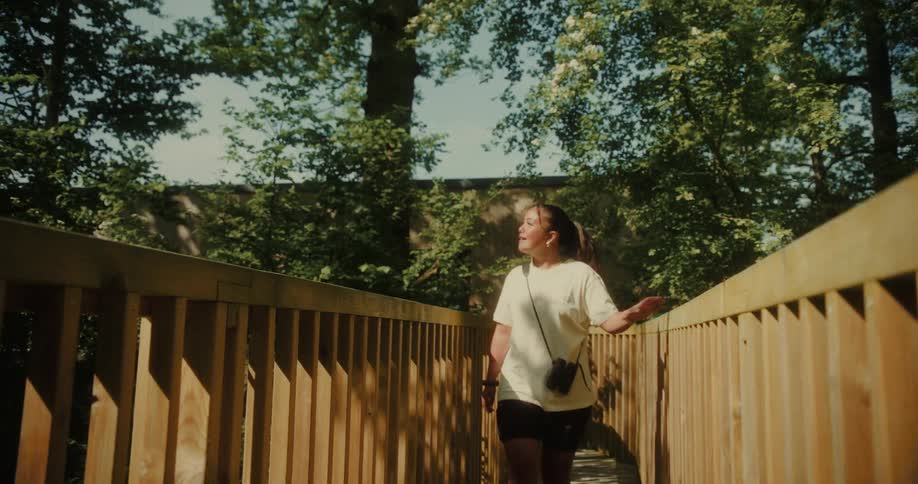 A young woman is walking on a pedestrian bridge at midday smiling and carrying a purse surrounded by trees