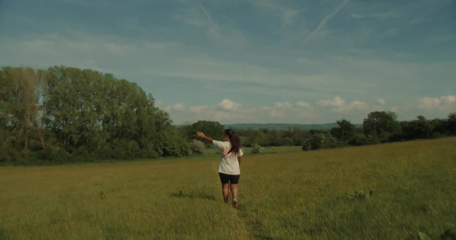 Woman in a white tshirt walking through a field with trees in the background