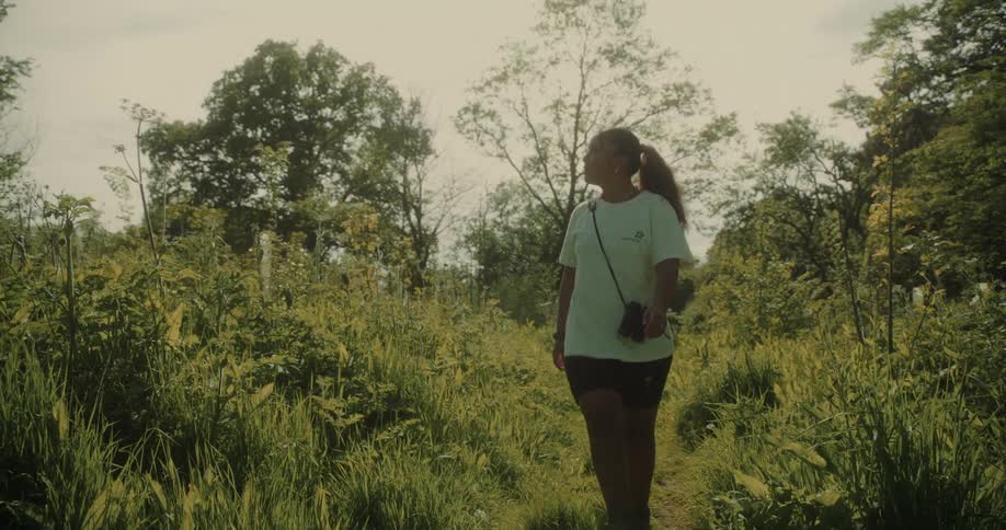 A young woman is walking on a path in the woods at midday while carrying a purse