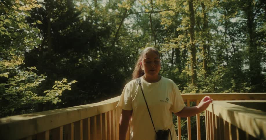 A young woman is walking on a pedestrian bridge at midday smiling and carrying a purse surrounded by trees