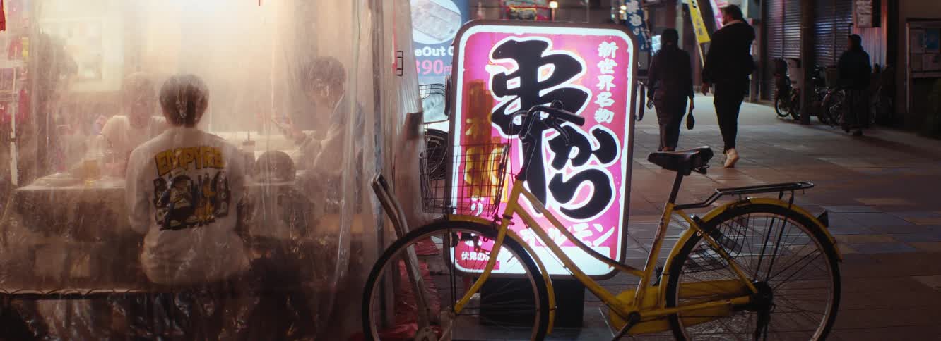 A yellow bicycle is parked in front of a brightly lit sign with japanese text on a busy street at night