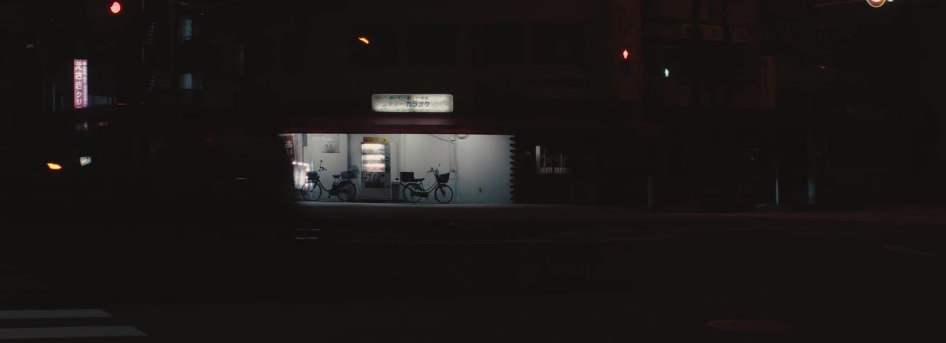 A small storefront with japanese signage sits along a dim street lit by a vending machine and flanked by two parked bicycles