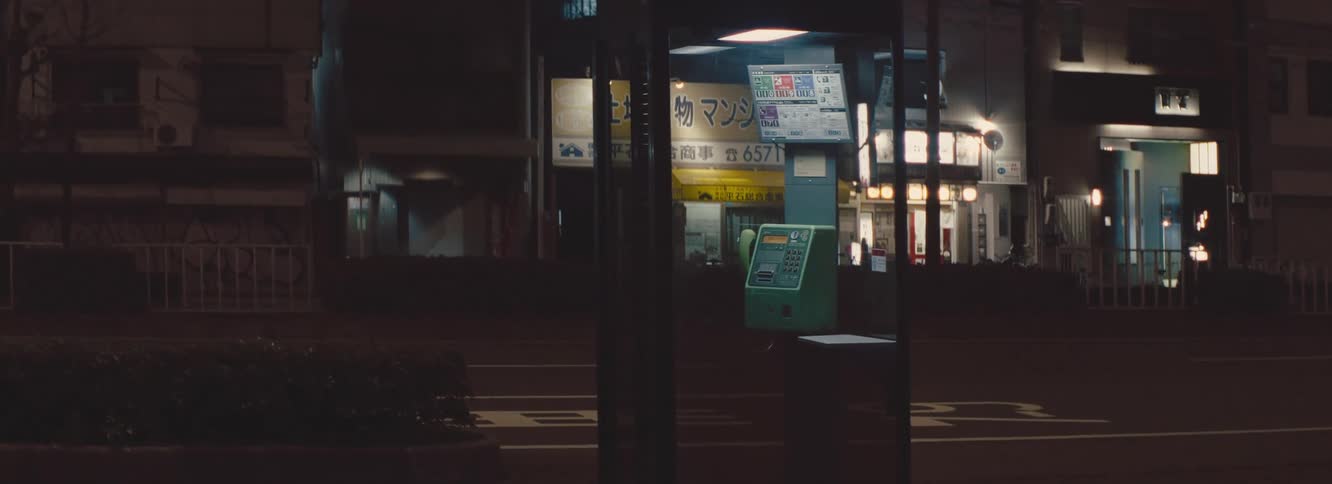A green payphone stands on a dimly lit street at night surrounded by glowing storefronts and signage