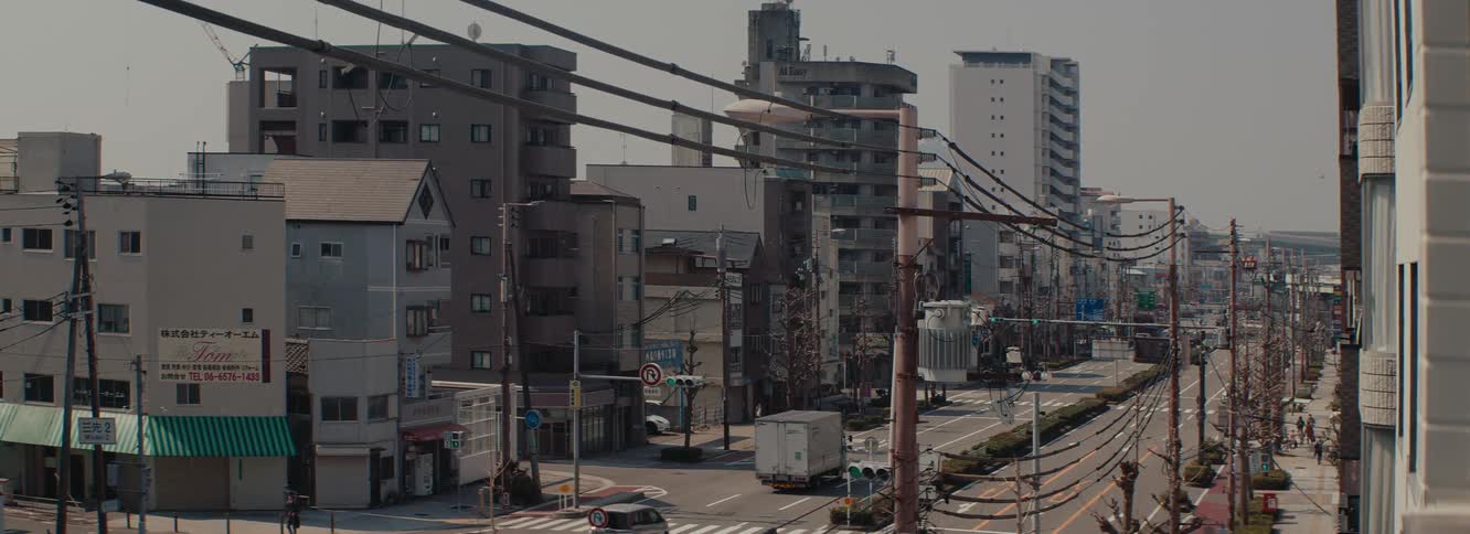 A city street lined with midrise buildings and overhead power lines with traffic lights and cars visible under an overcast sky in Japan