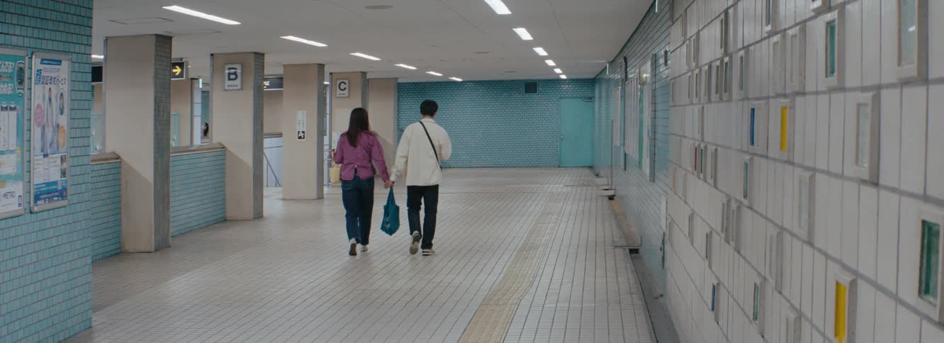 Two people walk down a well lit tiled subway or train station hallway passing signs for trains in both english and japanese the walls feature a distinctive blue mosaic tile pattern on one side and a textured light colored surface on the other