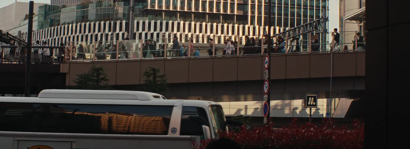 An exterior view of a modern building with a distinctive architectural design featuring horizontal white slats and large windows people can be seen walking on a walkway or bridge in front of the building with what appears to be green shrubbery