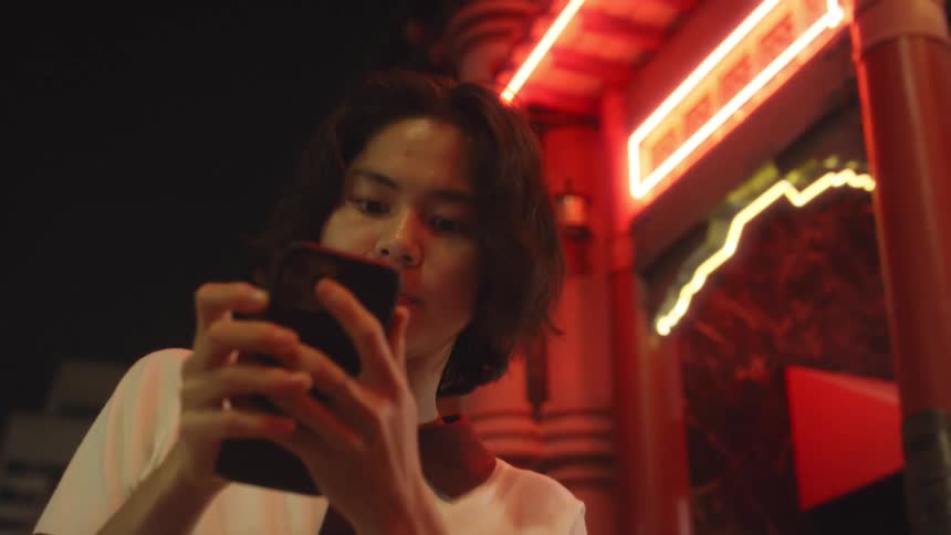 A person looks upward while holding a phone near a building lit by glowing red neon lights at night