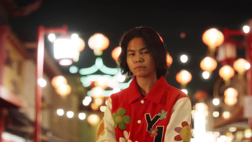 A young man in a red jacket decorated with flower patches stands on a lanternlit street at night surrounded by festive hanging lights
