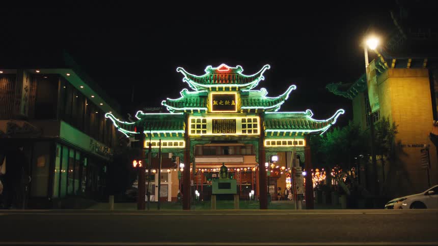A traditional chinese gate with glowing green neon outlines stands prominently at night backed by buildings and framed by bright signage and traffic lights