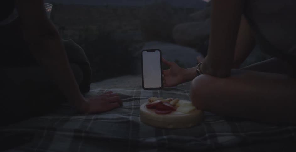 Two people sitting on a tablecloth with food and interacting with a smartphone