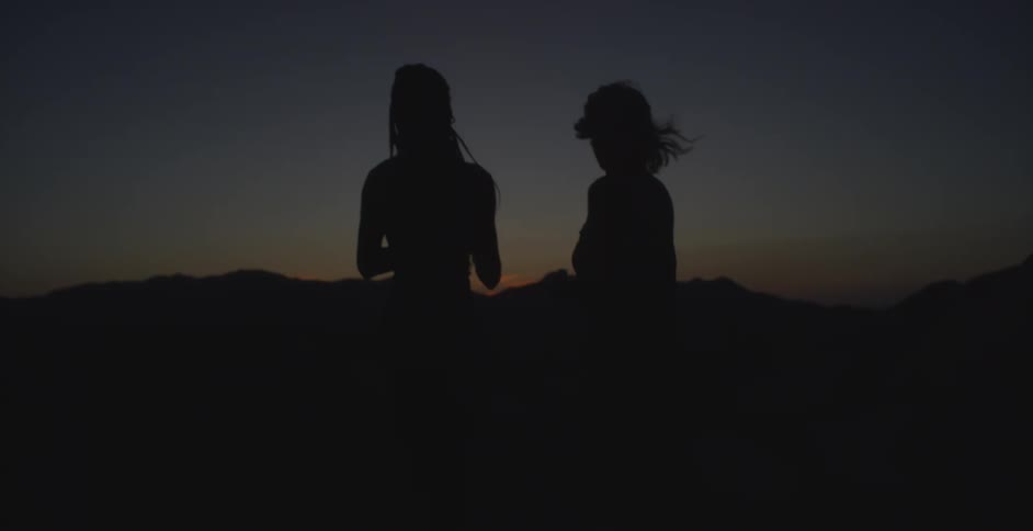 Two figures stand in silhouette against a dramatic sunset or sunrise with a wide expanse of sky and distant mountains one figure is holding up a phone capturing the moment