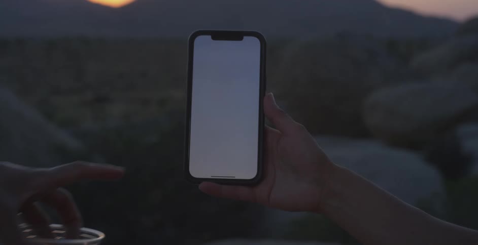 A hand holds a smartphone with a blank screen against twilightlit rocks and distant mountains