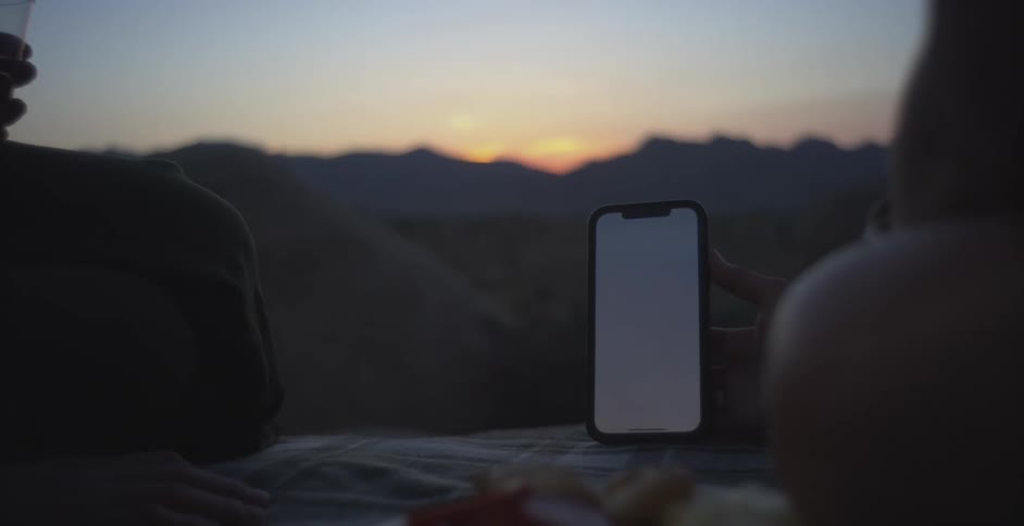 A phone is resting on a piece of cloth with two people sitting nearby during a mountain hike at sundown