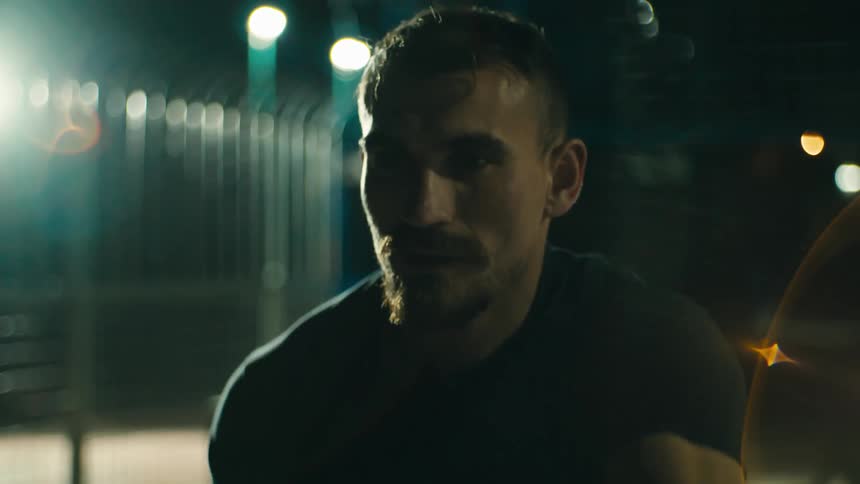 A person with a beard and mustache is shown in a closeup shot looking forward with bright lights in the background causing lens flares the individual appears to be sweating suggesting exertion or a warm environment