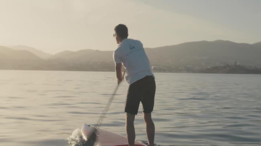 Two people paddleboard across a still lake at sunset with mountain silhouettes rising in the distance