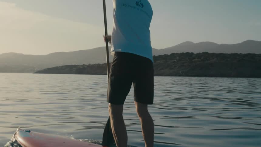Two men paddleboard across a calm sea towards a distant forested island under a clear sky