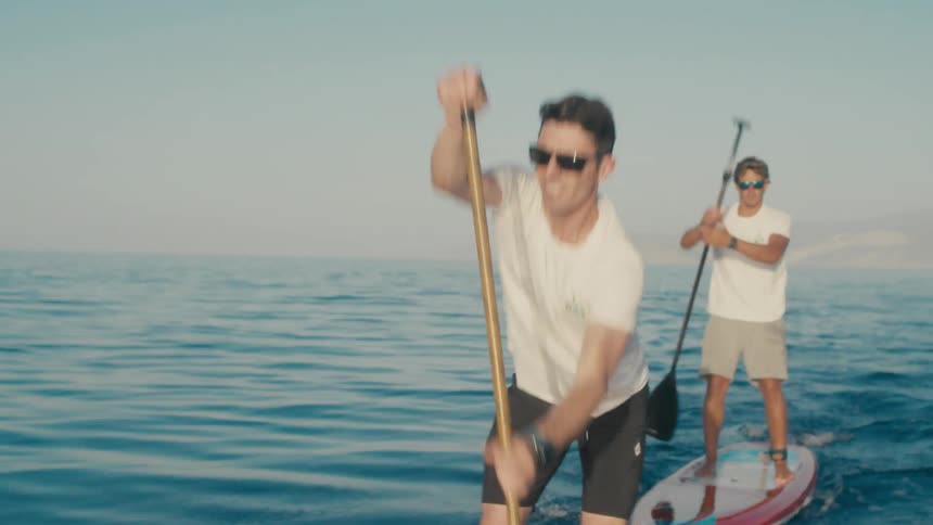 Two people paddleboard across a calm blue sea with a rocky island and hazy mountains in the distance under a clear sky