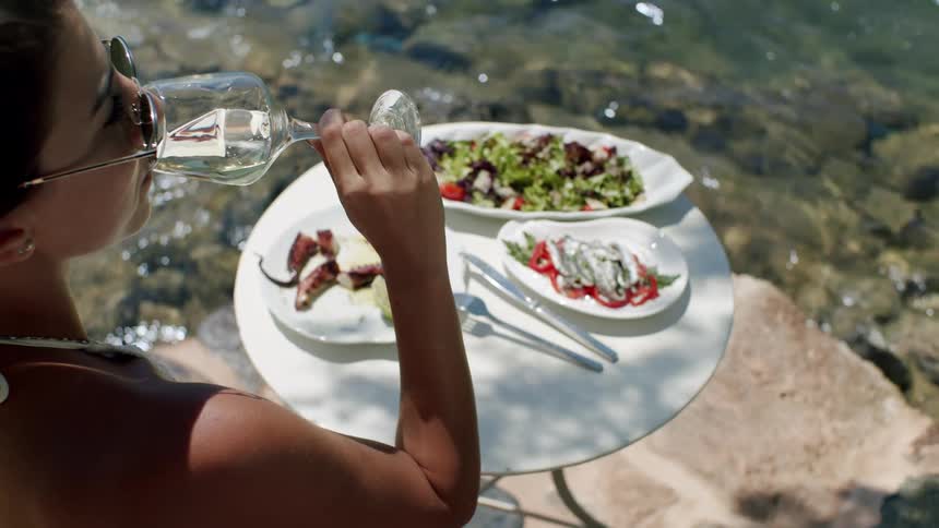 A person sits at a round table beside calm waters dining on white wine salad and seafood under soft outdoor light