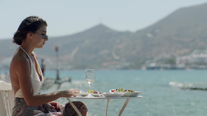A young woman is sitting by the beach on a chair under the sun eating from a meal served on a table with a glass of wine