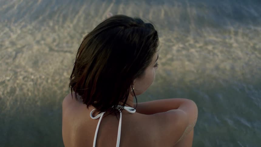 A woman in a swimsuit is squatting on the beach sand at midday staring at the water as waves crash on her feet