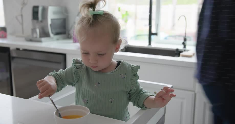 A toddler in a high chair stirs a bowl of food in a bright kitchen