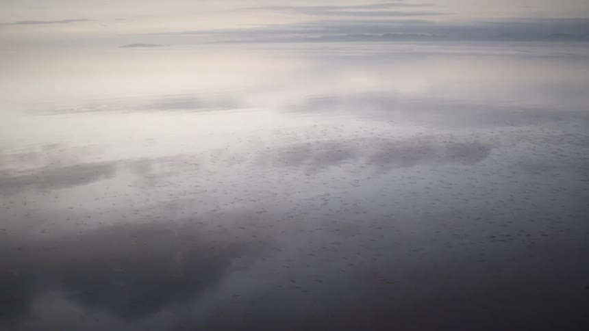 A calm lake mirrors a cloudy sky as mist drifts across the horizon in a serene quiet atmosphere