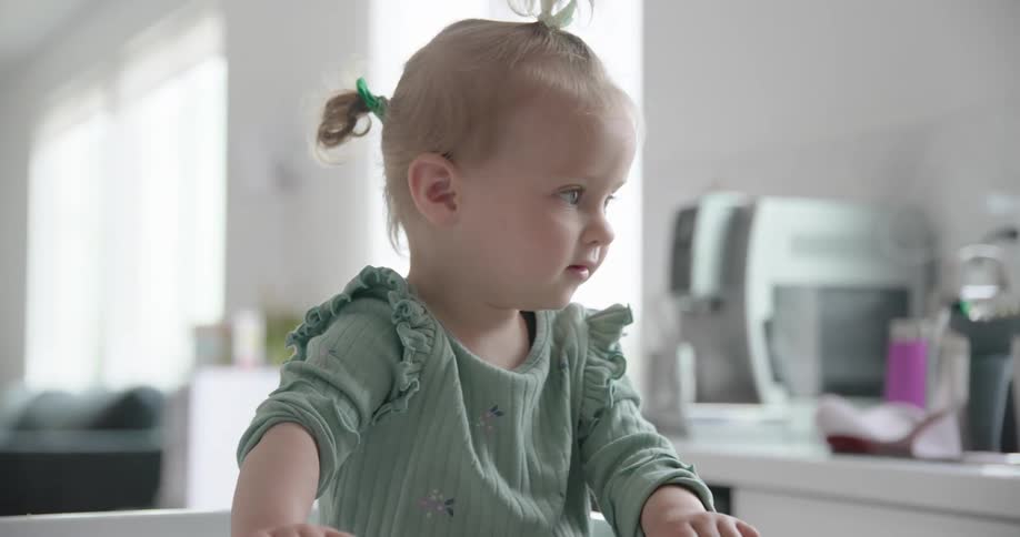 A toddler is in the kitchen sitting in her seat at midday