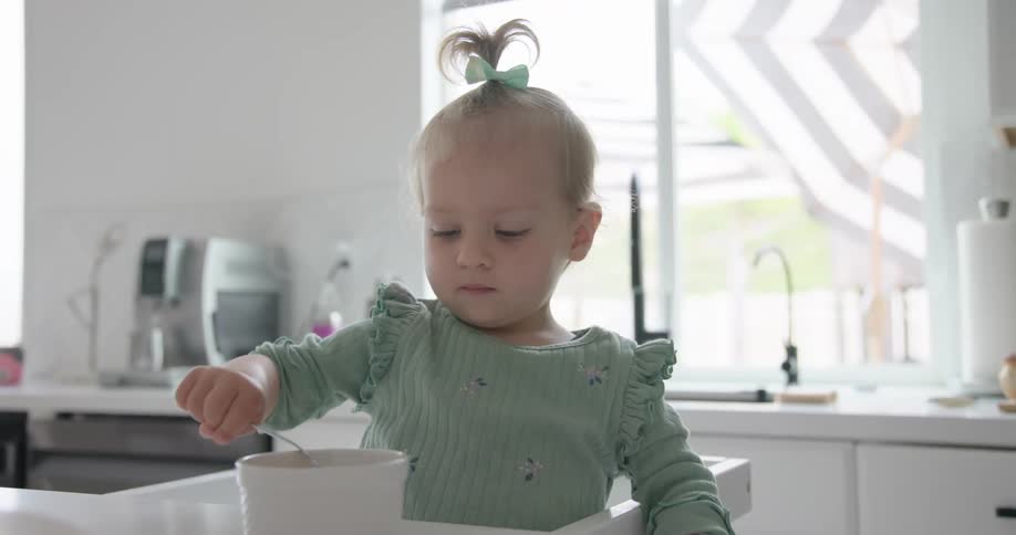 A young child with a green bow stirs a spoon in a white bowl while seated in a high chair inside a brightly lit kitchen