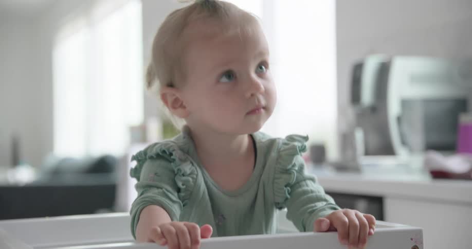 A young child in a green ruffled shirt stands at a sunlit kitchen counter focusing intently on a task