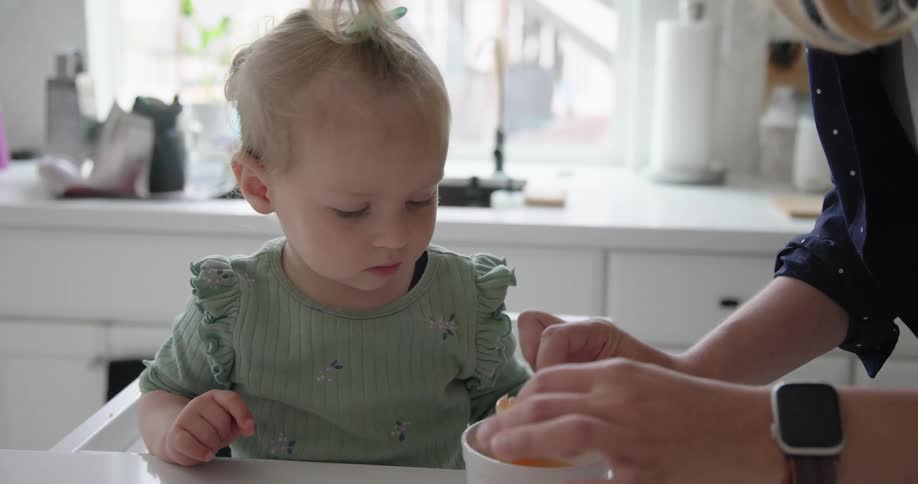 A woman in the kitchen cracks an egg onto a plate in front of her toddler at midday