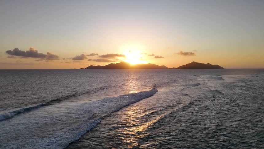 The sun sets behind a silhouetted island casting golden reflections across gentle ocean waves under a clear tranquil sky