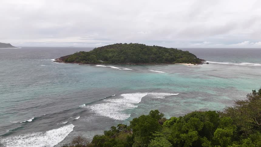 A small lush island surrounded by turquoise waters and waves is seen from a rocky green coastline under a cloudy sky