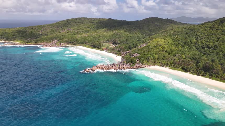 A stunning aerial shot captures a breathtaking tropical island where gentle waves wash ashore on white sand beaches surrounded by turquoise water and lush hills