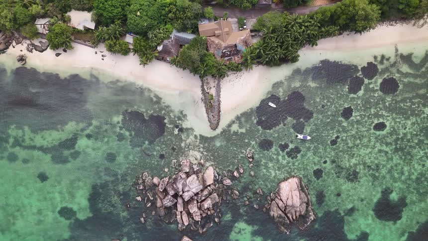 A tropical coastline curves around clear turquoise waters with rocky outcrops lush vegetation and scattered buildings along a sandy beach