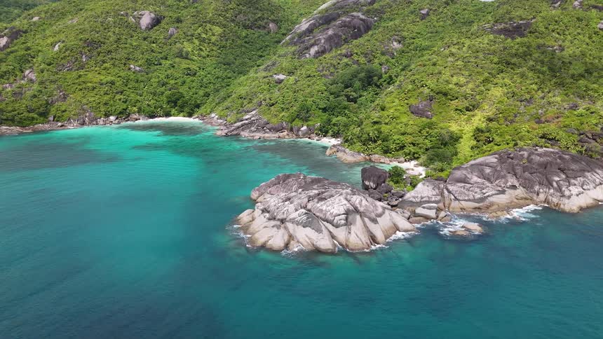 Turquoise waves lap against a rugged tropical coast lined with steep hills and rocky outcrops under a hazy sky