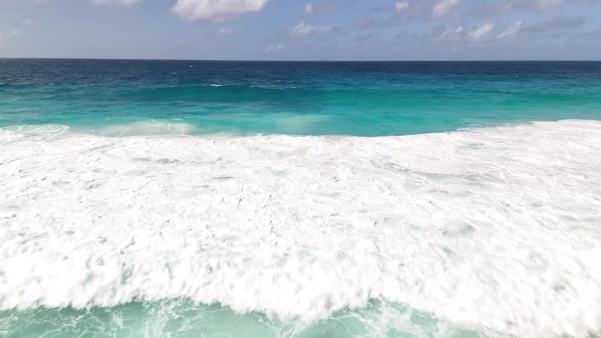 Waves crash onto a turquoise beach bordered by lush hills under a partly cloudy sky